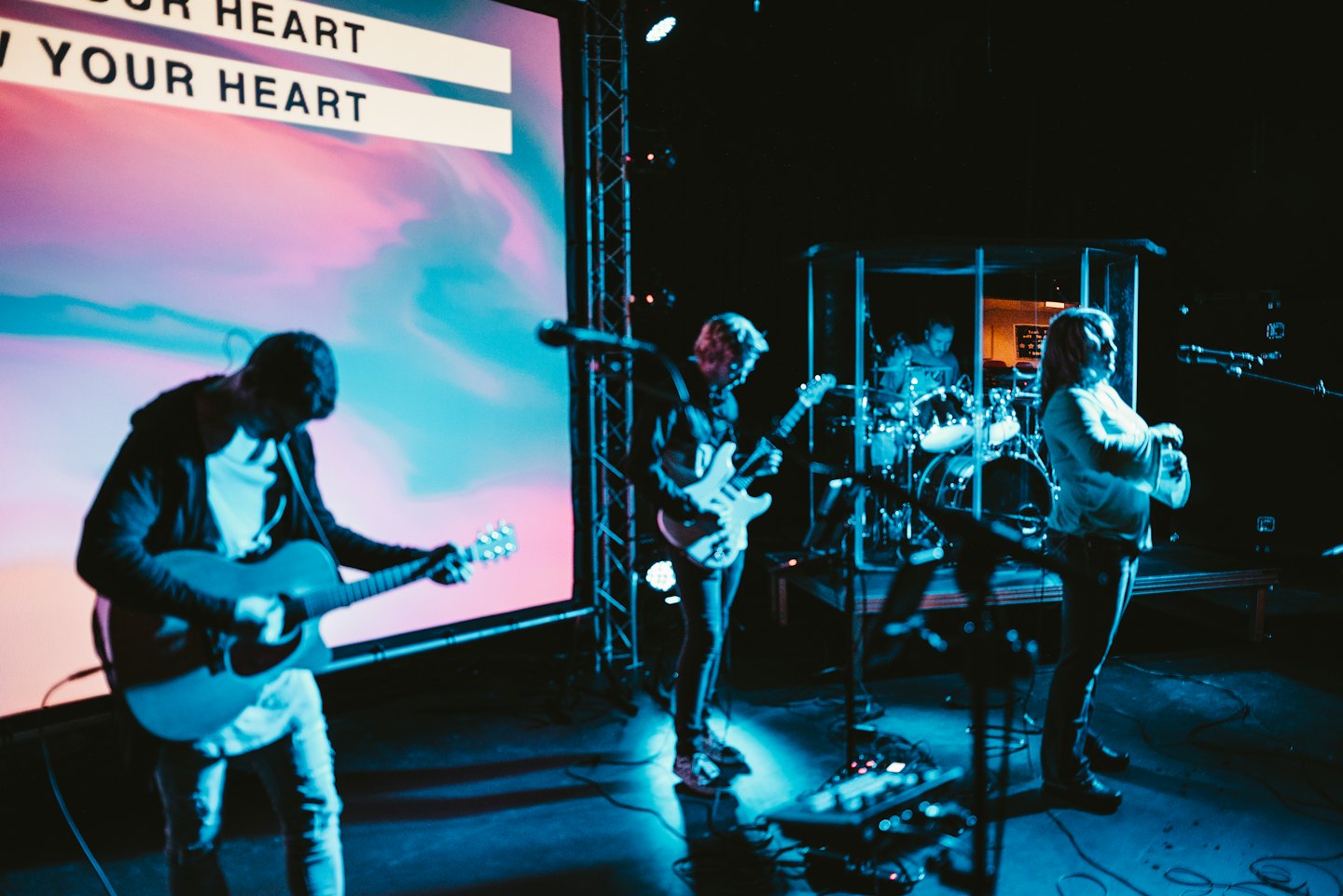 Una band dal vivo suona strumenti sul palco con testi visualizzati dietro.