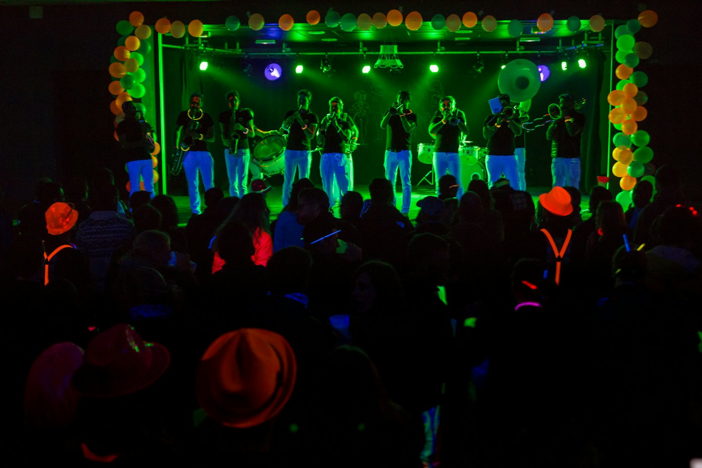 Musicisti suonano fiati e percussioni su un palco al neon con pubblico.